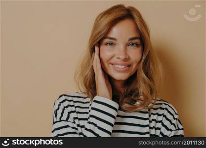 Portrait of good looking young woman touches face gently smiles toothily looks directly at camera dressed in casual striped jumper isolated over brown background has healthy well cared skin.. Portrait of good looking young woman touches face gently smiles toothily looks directly at camera