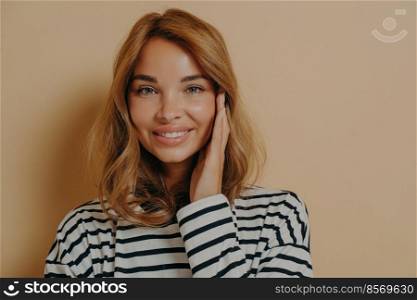 Portrait of good looking young woman touches face gently smiles toothily looks directly at camera dressed in casual striped jumper isolated over brown background has healthy well cared skin.. Portrait of good looking young woman touches face gently smiles toothily looks directly at camera