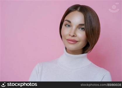 Portrait of good looking young woman with dark hair, minimal makeup, wears casual white turtleneck, going to have walk, poses against rosy studio wall, blank space for your advertising content.