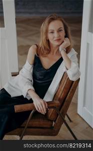 Portrait of gorgeous redhead woman in black evening dress and white shirt. Stylish confident mid adult lady is sitting in an arm chair at home. Happy european woman is going off to a party.. Portrait of gorgeous confident mid adult lady sitting in an arm chair. Woman in black evening dress.