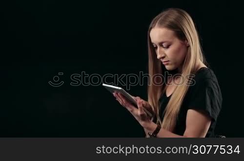 Portrait of gorgeous woman working on touchpad against black background. Attractive blonde female surfing the net with tablet pc and looking at screen. Confused girl searching web with digital tablet.