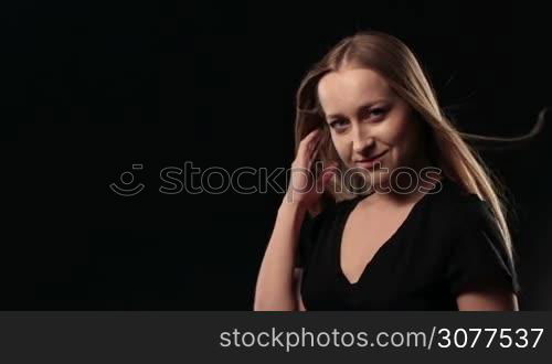 Portrait of gorgeous young blonde woman with amazing full hair and deep blue eyes posing at studio on black background. Sensual female with fluttering hair in the wind looking at camera and smiling. Slow motion.