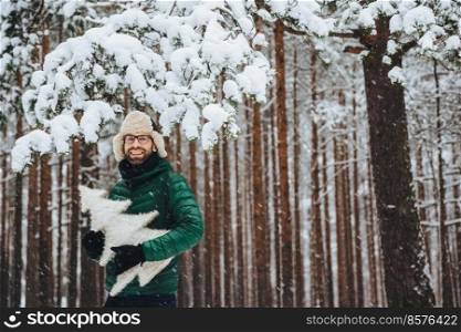 Portrait of handsome bearded male wears warm fur cap with ear flaps and green anorak, holds artificial fir tree, poses against high trees covered with snow, spends free time in winter forest