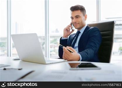 Portrait of Handsome businessman sitting talking on smartphone and using laptop at workplace. Confident Smart Man Manager smiling working for business and financial near window in room at office