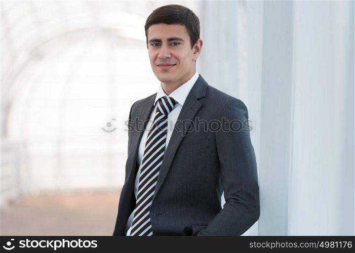 Portrait of handsome confident young businessman standing arms in pockets, smiling happy, looking at camera.