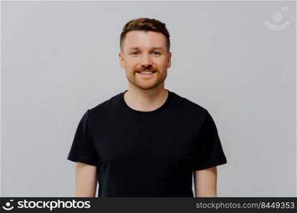 Portrait of handsome European man with stubble dressed in casual black t shirt smiles happily poses against blue background expresses positive emotions. Carefree glad male model in good mood