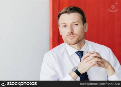 Portrait of handsome male employee has stubble, holds hands together, looks thoughtfully at camera, contemplates about business project, poses against red and white background with copy space