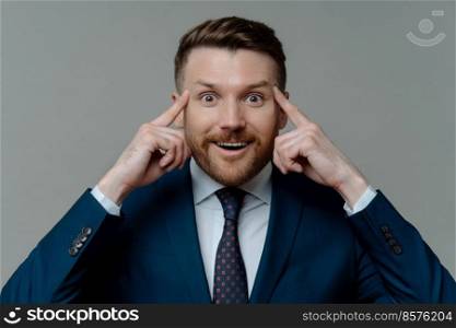 Portrait of handsome male entrepreneur keeps fingers on temples looks with happy surprised expression at camera tries to recall information isolated over grey background. Experienced businessman