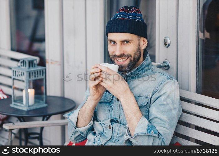 Portrait of handsome male with cheerful expression, enjoys spare time, drinks coffee or tea, wears hat and denim jacket, being in good mood. Attractive man has thick beard and mustache poses indoor