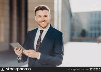 Portrait of handsome male with positive expression, updates software on tablet computer, connected to wireless internet, shares files during remote job, stands outdoor, reads company s news.