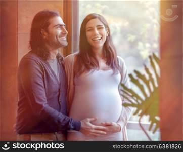Portrait of handsome man touching tummy of his pregnant wife, standing near window in bright sunlight, happy pregnancy time