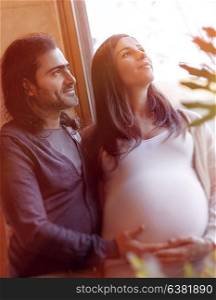 Portrait of handsome man touching tummy of his pregnant wife, standing near window in bright sunlight and with hope looking in future