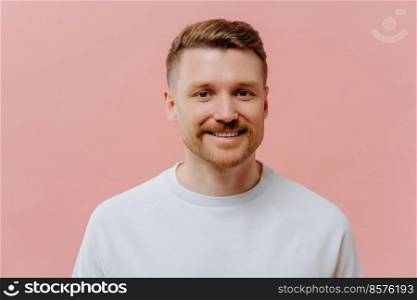 Portrait of handsome pleasant unshaven redhead man expressing positiveness while looking at camera with smile, happy guy in white tshirt feeling joyful while posing against pink studio background.. Young positive man smiling at camera in pink studio background