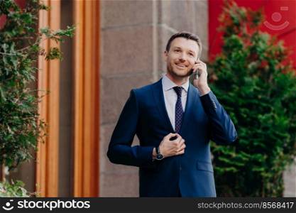 Portrait of handsome satisfied successful businessman talks on cell phone, discusses banking transactions, dressed in corporate suit, has positive expression. Business and technology concept