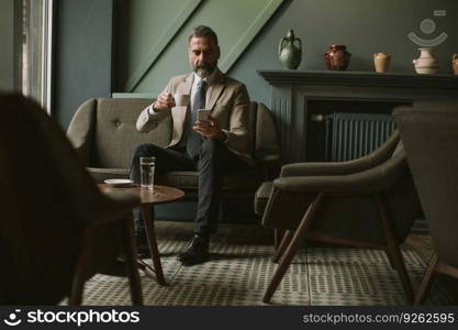 Portrait of handsome senior businessman drinking coffee and using mobile phone in lobby