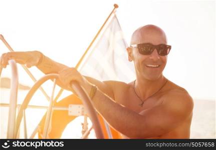 Portrait of handsome shirtless sailor behind helm of sailboat in bright sunny day, enjoying active summer holidays, happy traveling along sea