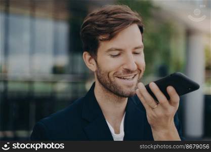 Portrait of handsome smiling bearded office worker talking on speakerphone, recording audio message, using virtual digital voice assistant, standing outside of mirrored business center building. Handsome smiling neat bearded office worker talking on speakerphone standing outside