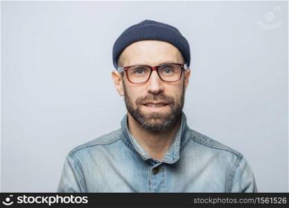 Portrait of handsome stylish male journalist wears eyewear, fashionable hat and denim jacket, poses against white studio background. Satisfied man with stubble has blue eyes and pleasant appearance
