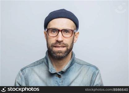Portrait of handsome stylish male journalist wears eyewear, fashionable hat and denim jacket, poses against white studio background. Satisfied man with stubble has blue eyes and pleasant appearance