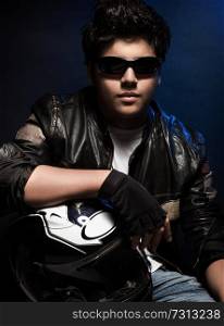 Portrait of handsome stylish teen boy posing in the studio over dark blue background, wearing bikers outfit and holding helmet in hands
