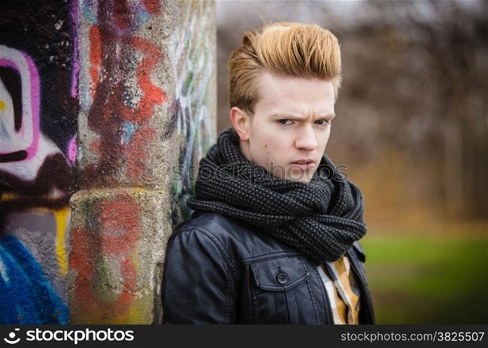 Portrait of handsome trendy man outdoor in city setting, male model wearing winter clothes black jacket and scarf against colorful graffiti wall