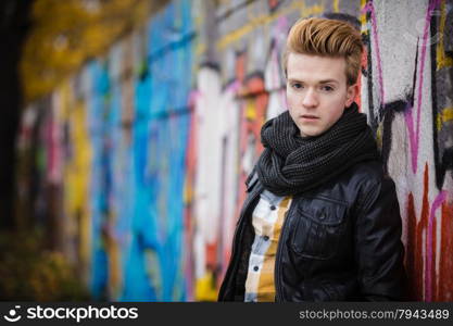 Portrait of handsome trendy man outdoor in city setting, male model wearing winter clothes black jacket and scarf against colorful graffiti wall