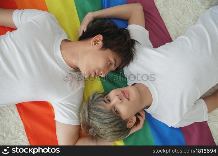 Portrait of handsome young gay couple lying on the rainbow LGBT flag and looking at each other and camera at bedroom