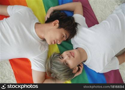 Portrait of handsome young gay couple lying on the rainbow LGBT flag and looking at each other and camera at bedroom