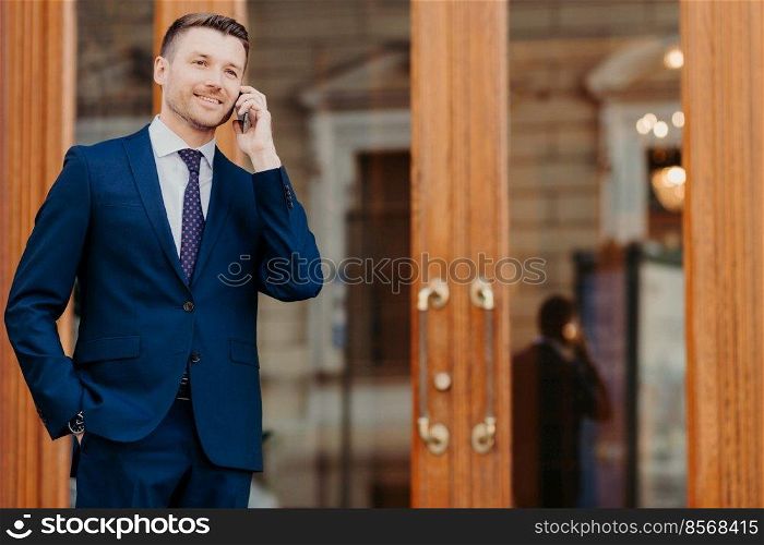 Portrait of handsome young male boss dressed in elegant expensive suit has mobile phone conversation, stands near door of cafe, rests after meeting with partners. People, business and technology