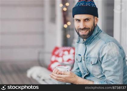 Portrait of handsome young smiling male enjoys spare time, holds mug of coffee, being deep in thoughts, has coffee break during shining warm day, focused on something. Pleased bearded stylish man