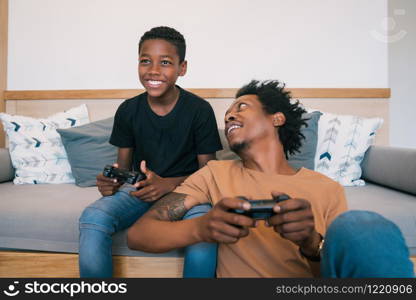 Portrait of happy African American father and son sitting in sofa couch and playing console video games together at home. Family and technology concept.