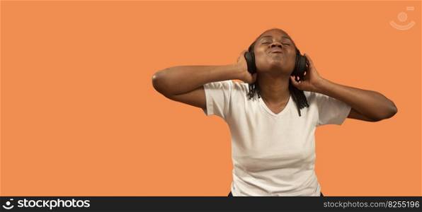 Portrait of happy African young woman listening to music with headphones, eyes closed, head up and holding headphones with hands, dressed in white t-shirt against orange background with copy space. Portrait of African young woman listening to music with headphones, eyes closed, head up and holding headphones with hands against orange background