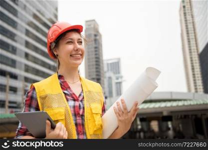 Portrait of happy American young woman engineer with safety helmet, paper blueprint, digital tablet celebrate project site at town complete. Real estate construction in city.