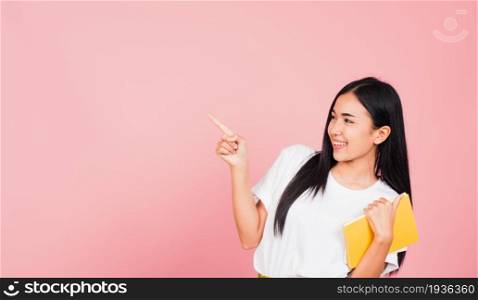 Portrait of happy Asian beautiful young woman confident smiling holding orange book open pointing finger to side copy space, studio shot isolated on pink background, education concept