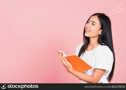 Portrait of happy Asian beautiful young woman confident smiling standing holding orange book open or diary for reading, studio shot isolated on pink background, with copy space, education concept