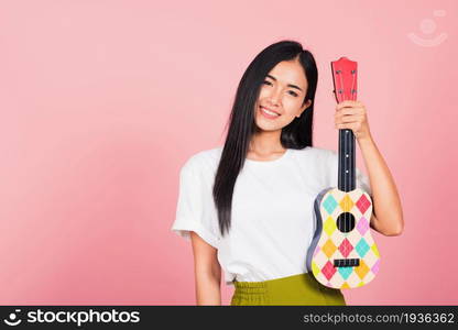 Portrait of happy Asian beautiful young woman teen confident smiling face hold acoustic Ukulele guitar, female playing Hawaiian small guitar, studio shot isolated on pink background, with copy space