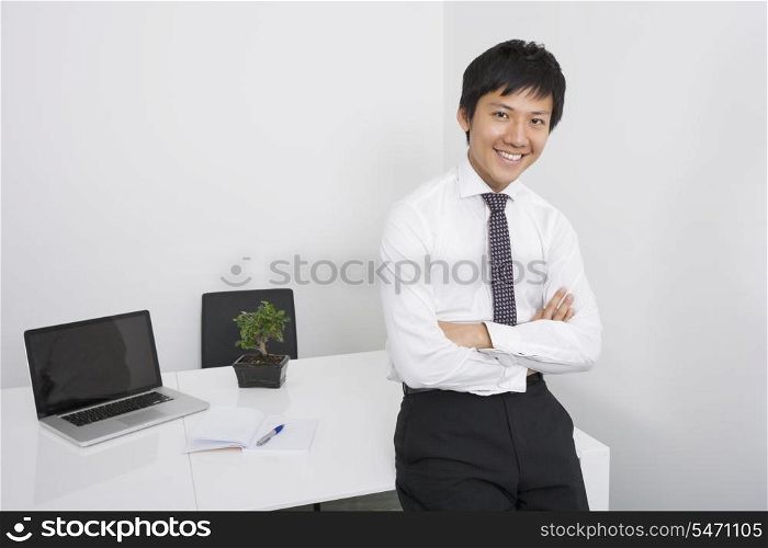 Portrait of happy Asian businessman with arms crossed leaning on office desk