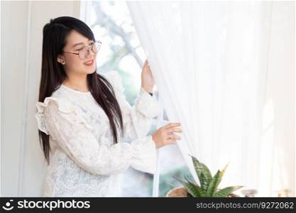 Portrait of Happy Attractive asian people cute woman wearing white lace shirt with the curtains on the window in the room felt like relaxing in house like the background