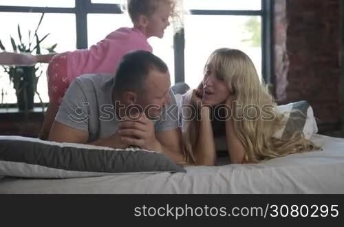 Portrait of happy attractive family with lovely little daughter lying on bed and smiling. Carefree parents with cute girl spending leisure together at home while relaxing at bed in morning. Slow motion. Dolly shot.