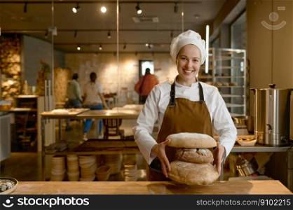 Portrait of happy baker holding stack of freshly baked bread. Young female chef proud of making natural organic pastry standing at table and looking at camera. Young happy baker holding stack of freshly baked bread looking at camera