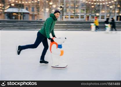 Portrait of happy bearded man wears fashionable clothes uses skate aid for keeping balance on ice, enjoys weekends in circle of close friends, poses at camera. People, leisure, lifestyle concept