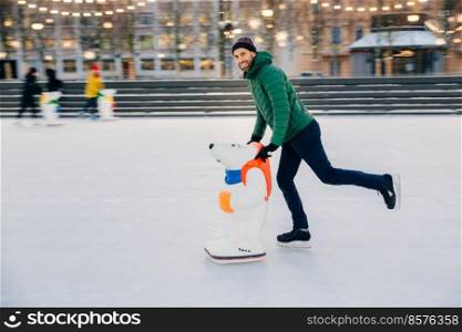 Portrait of happy bearded man wears fashionable clothes uses skate aid for keeping balance on ice, enjoys weekends in circle of close friends, poses at camera. People, leisure, lifestyle concept