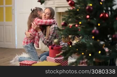 Portrait of happy beautiful mother and cute daughter embracing in Christmas decorated interior at home. Joyful little girl hugging her affectionate mom tenderly and both looking at camera with toothy happy smiles during winter holidays. Dolly shot.