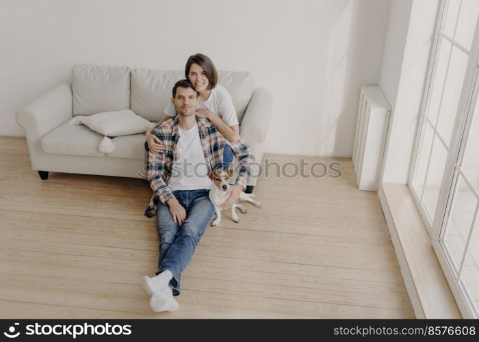 Portrait of happy brunette young woman embraces with love her husband who sits on floor with pedigree dog, pose in empty spacious room, move in new apartment after marriage, start family life