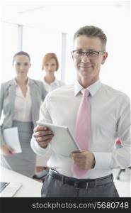 Portrait of happy businessman holding digital tablet with female colleagues in background at office