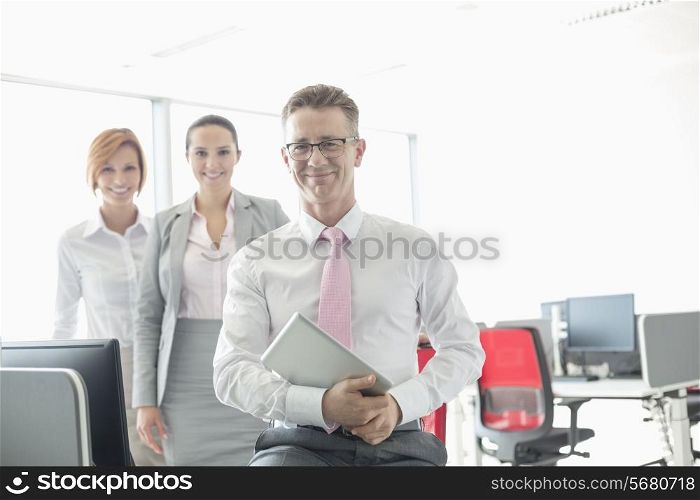 Portrait of happy businessman holding tablet PC with female colleagues in background at office