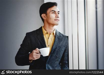 Portrait of Happy Businessman Standing by the Window in Office. Looking Away and Smiling. Dreaming for Success. Drinking Hot Coffee