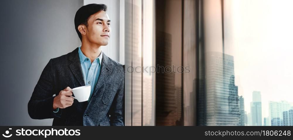 Portrait of Happy Businessman Standing by the Window in Office. Looking Away and Smiling. Dreaming for Success. Drinking Hot Coffee