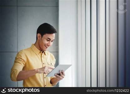 Portrait of Happy Businessman Standing by the Window in Office. Using Digital Tablet and Smiling