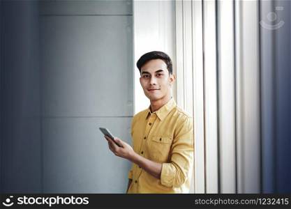 Portrait of Happy Businessman Standing by the Window in Office. Using Smartphone and Smiling. Looking at Camera
